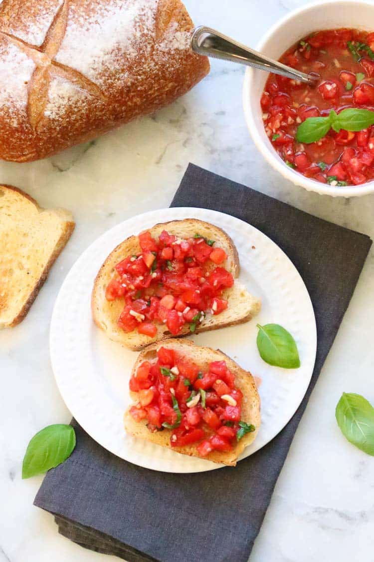 wide overhead shot of bruschetta on toast with bowl of bruschetta and loaf of french bread nearby