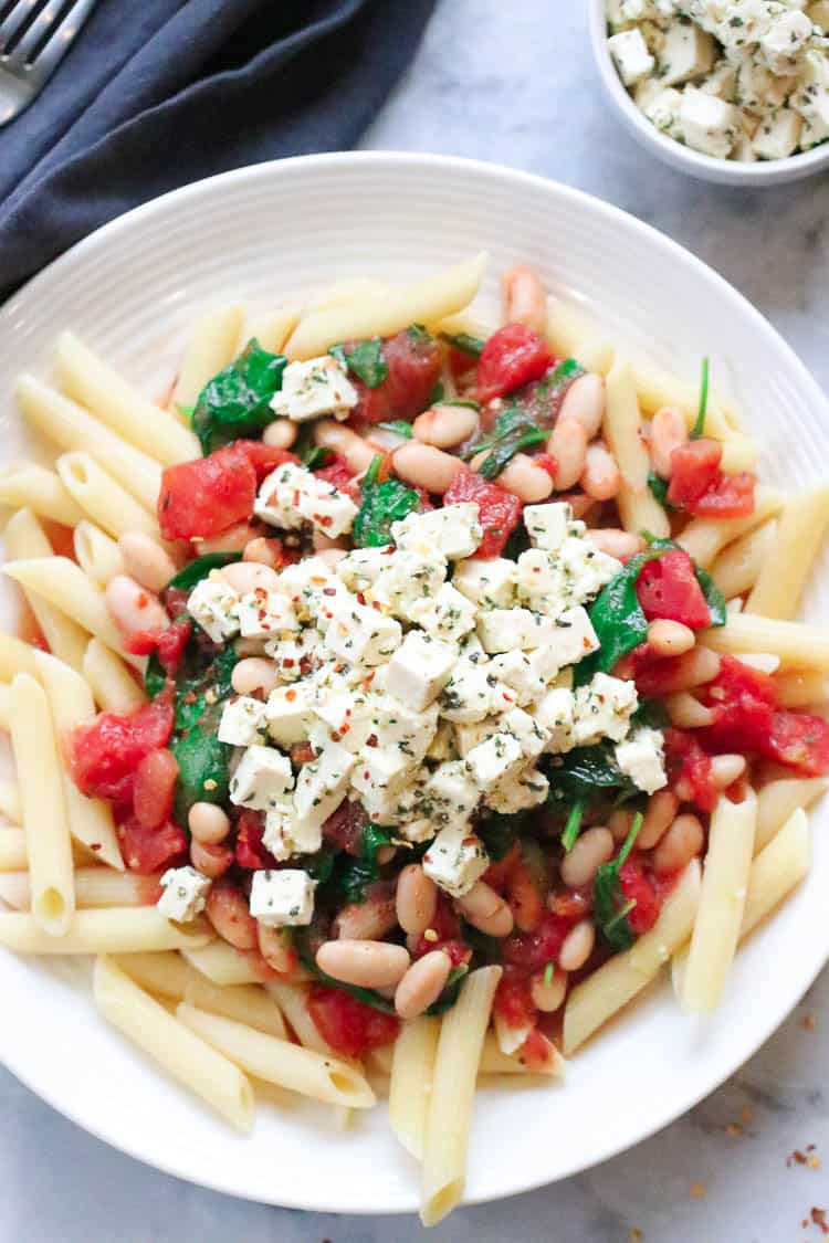 overhead closeup shot of pasta with Cannellini beans, spinach, Italian tomatoes and vegan feta on a white plate with ramekin beside