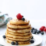 side shot of vegan pancakes stacked on a plate topped with fresh berries and dripping syrup