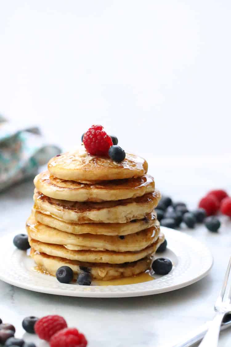 side shot of vegan buttermilk pancakes stacked tall on a white plate with utensils nearby and garnished with fresh blueberries and raspberries