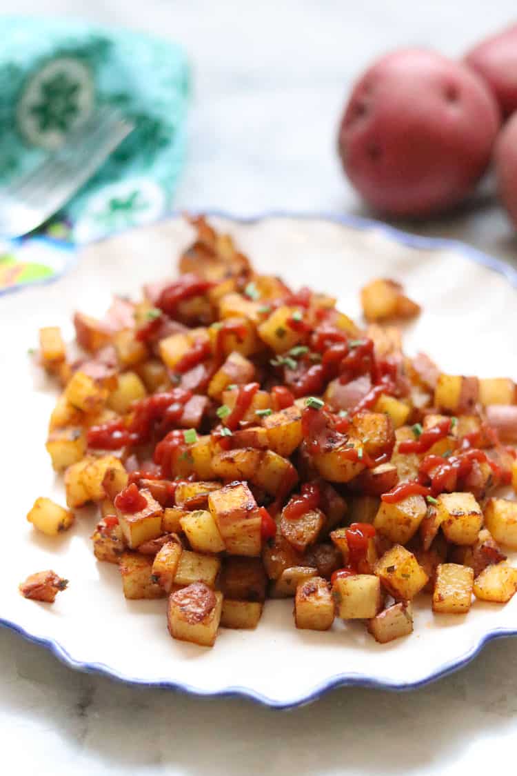 side shot of diced baked breakfast potatoes on a white plate with red potatoes and napkin in the background