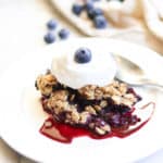 side shot of a serving of blueberry crisp on a white plate topped with whipped cream