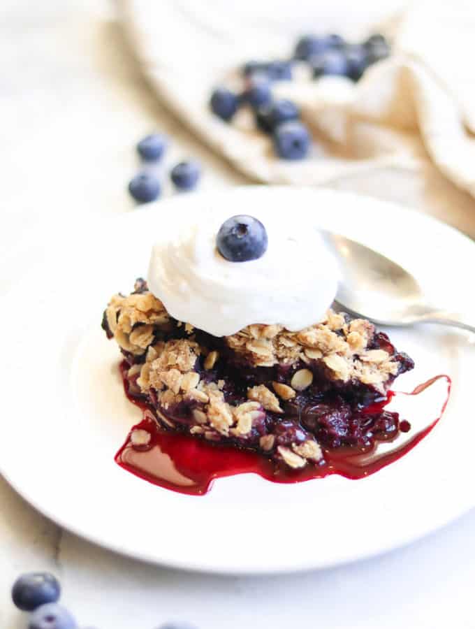 side shot of a serving of blueberry crisp on a white plate topped with whipped cream