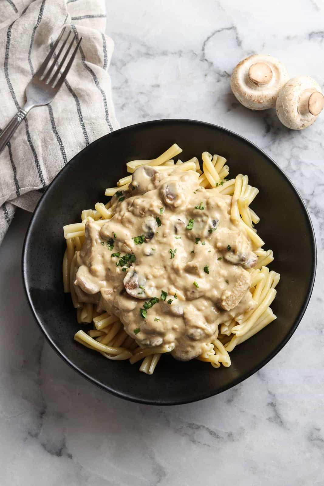 overhead photo of stroganoff on a bed of pasta in a black pasta bowl with a garnish of chopped green herbs