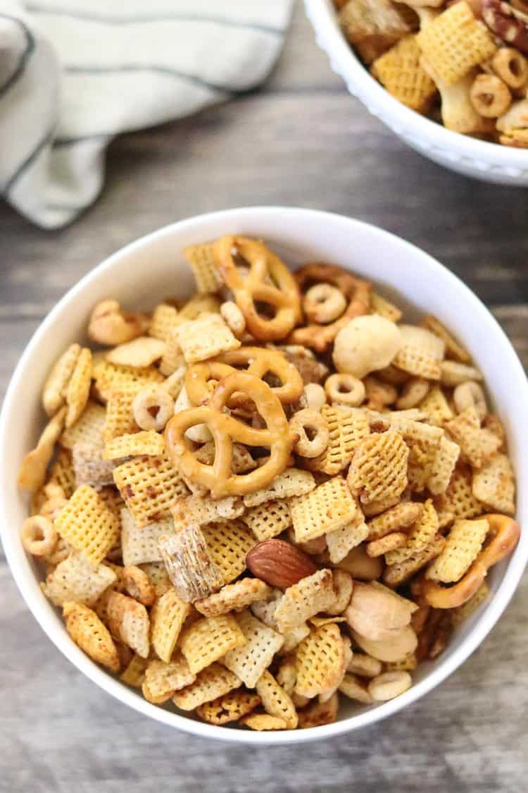 overhead shot of chex mix in white bowl on brown wooden background with napkin beside