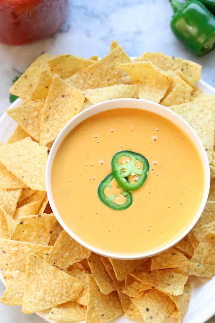 vegan cheese sauce in a white bowl surrounded by nacho chips and decorated with sliced jalapeño