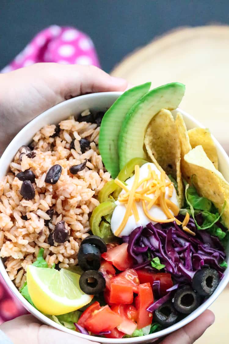 Two Hands holding white bowl with Mexican Rice and Black Beans and a variety of toppings.