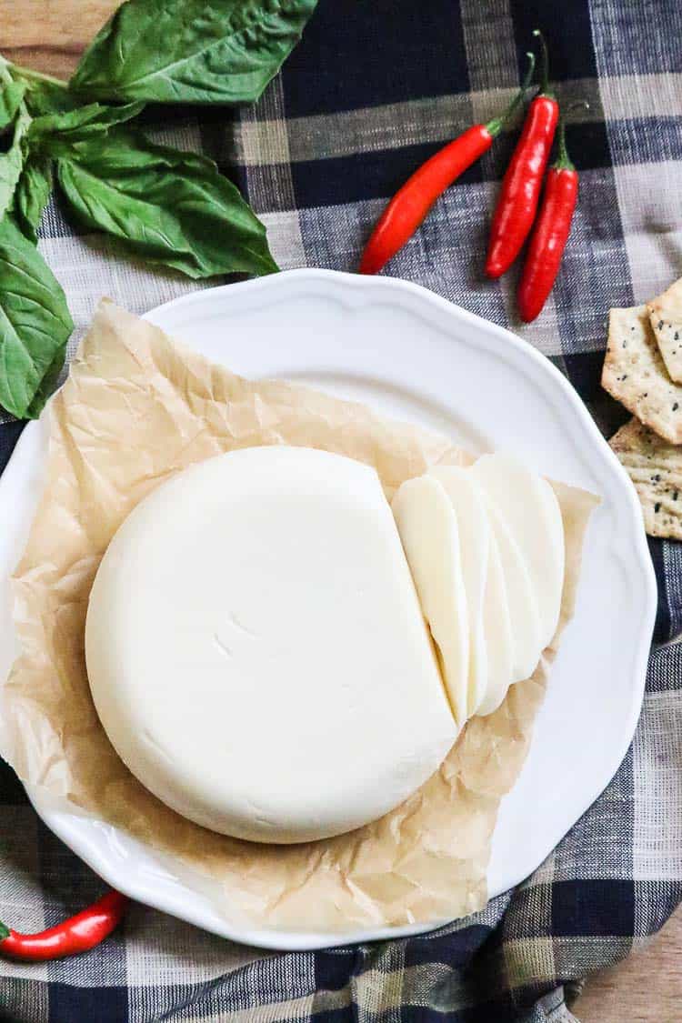 vegan mozzarella cheese block sliced on a white plate with red peppers and basil beside.