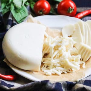 side view of vegan mozzarella cheese sliced and grated on a white plate with a plaid napkin underneath and basil, tomatoes and peppers beside