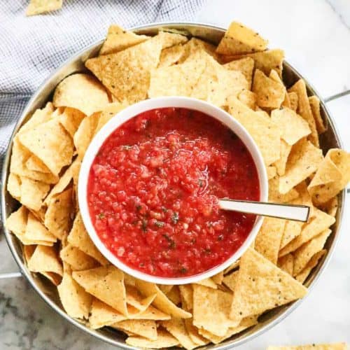 overhead view of salsa in a white bowl with chips in a silver dish surrounding it