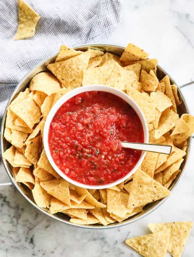 overhead view of salsa in a white bowl with chips in a silver dish surrounding it