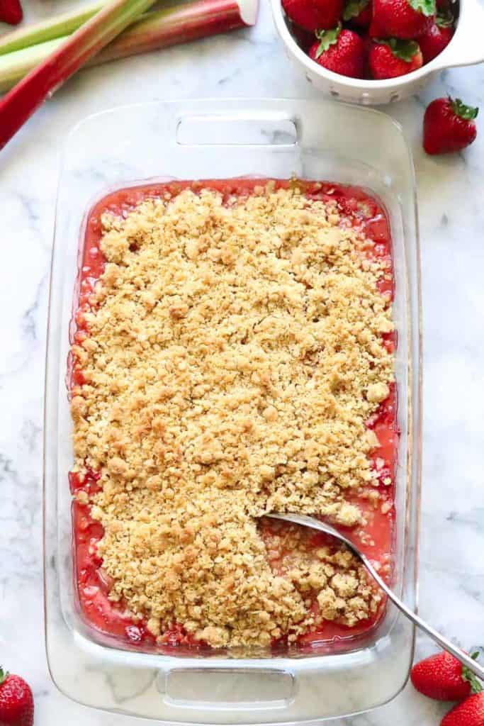 Casserole dish filled with strawberry rhubarb crisp and rhubarb stalks and strawberries beside.
