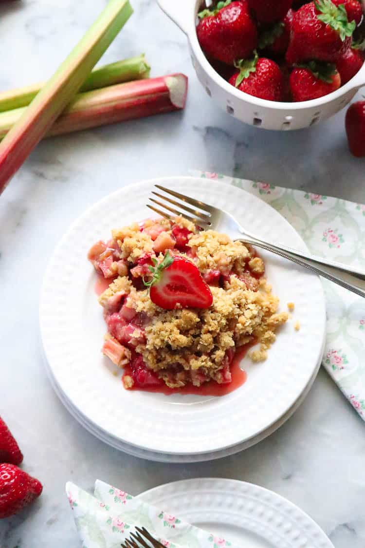 Vegan Strawberry Rhubarb Crisp on a plate with a sliced strawberry on top and stacked plates beside.