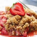 Closeup shot of Strawberry Rhubarb Crisp on a plate with a fork beside and a sliced strawberry on top