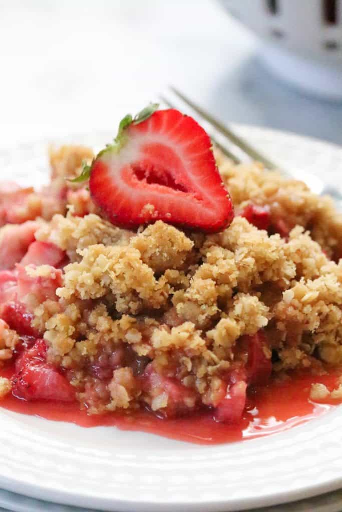 Closeup shot of Strawberry Rhubarb Crisp on a plate with a fork beside and a sliced strawberry on top