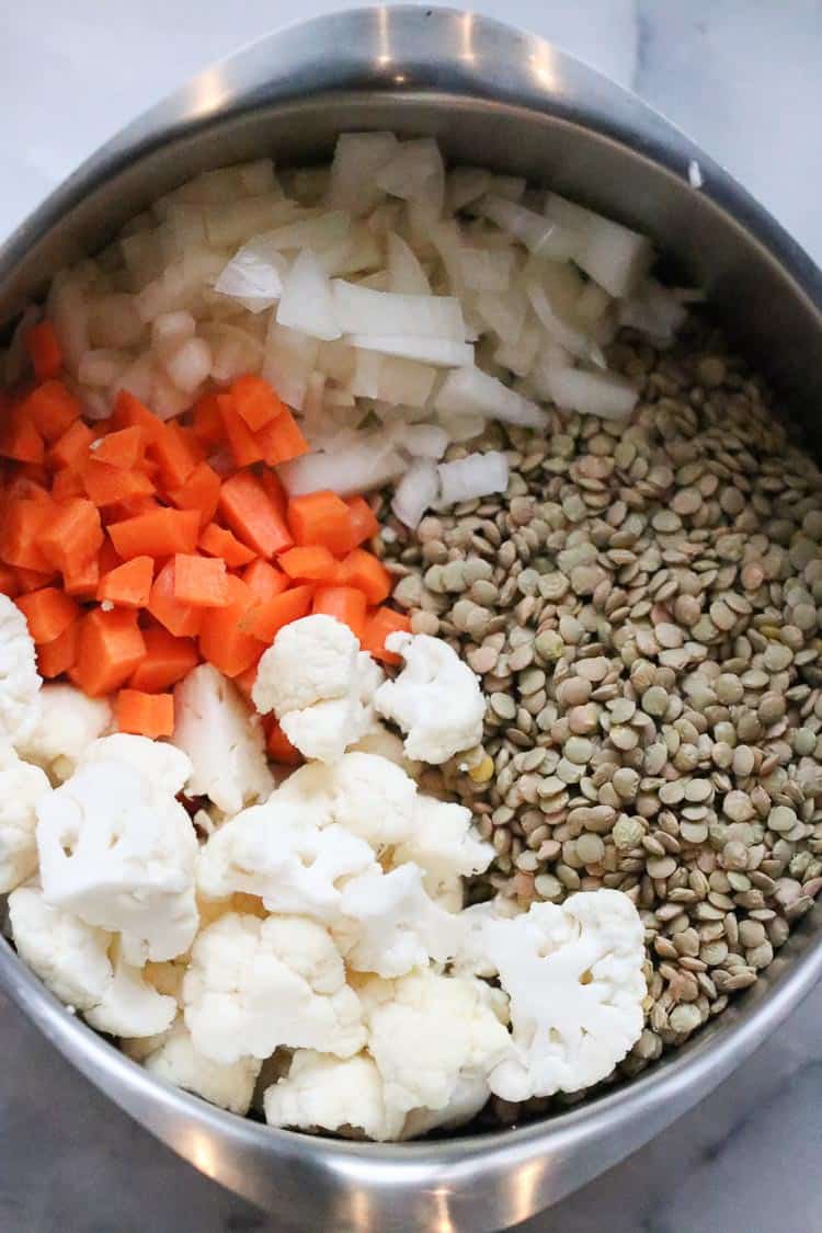 overhead shot of brown lentils, chopped carrots, cauliflower, and diced onion in a med sized silver soup pot
