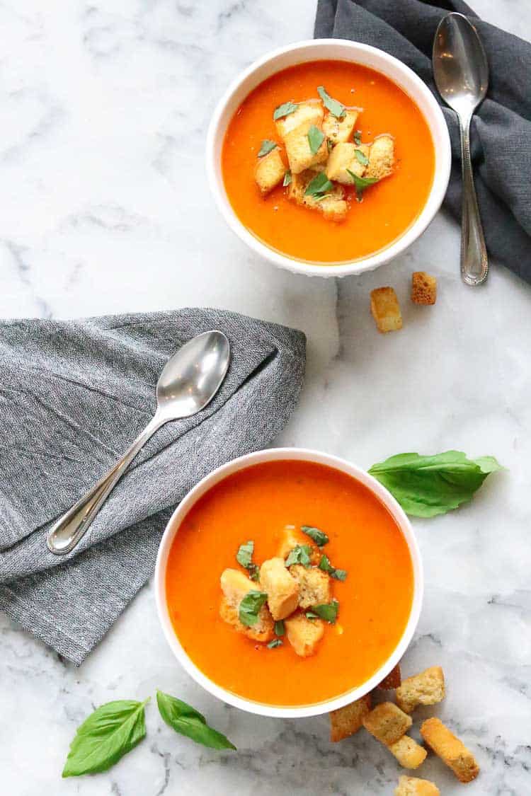 overhead shot of two white bowls filled with vegan tomato soup with grey napkins beside and croutons and basil strewn about