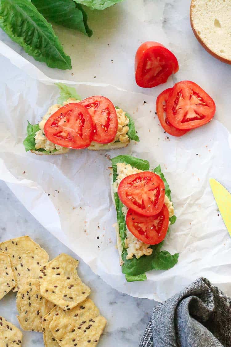 wide overhead shot of vegan chickpea salad sandwiches with lettuce, cut tomato and chips nearby