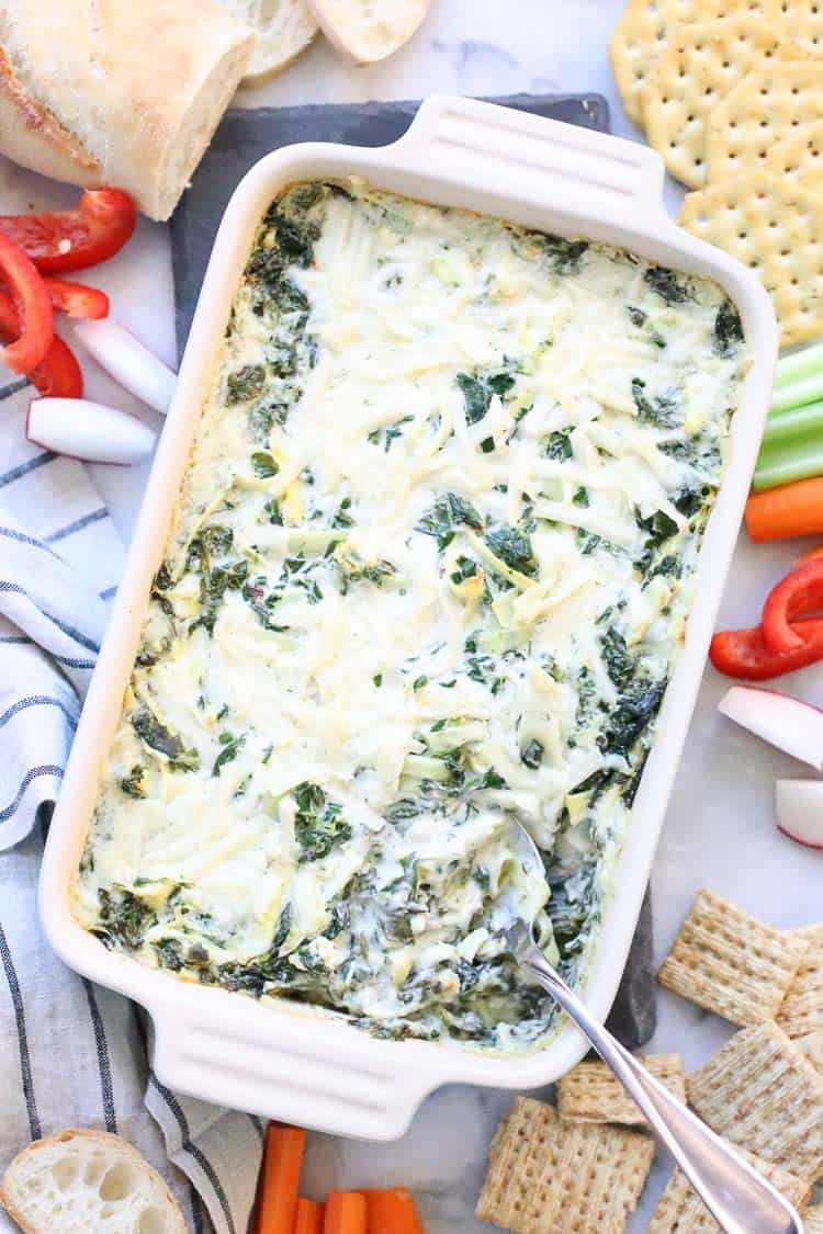 overhead shot of vegan spinach artichoke dip in casserole dish with crackers, veggies and bread surrounding