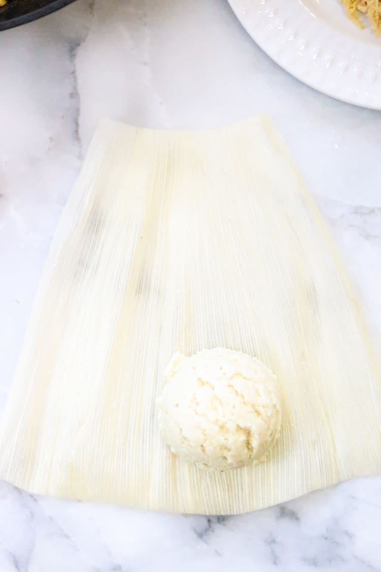 overhead shot of vegan tamale creation with corn husk laid on white background and a scoop of tamale dough on top