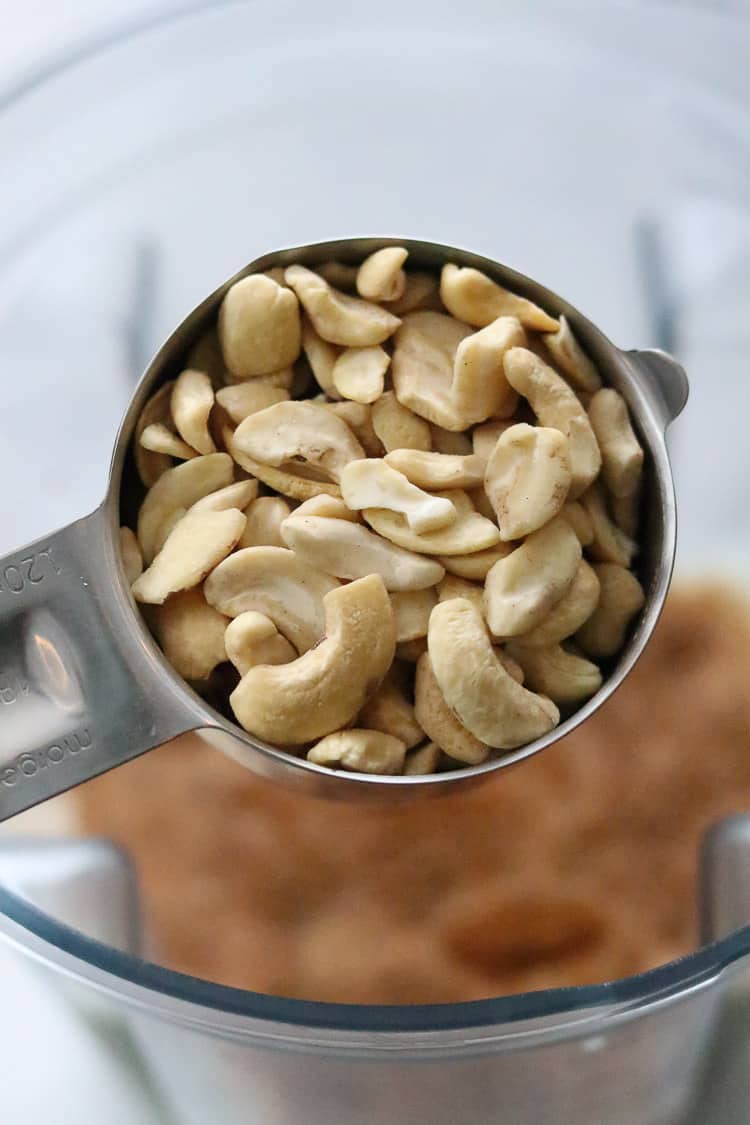 overhead shot of measuring cup full of cashews held over blender full of ingredients for vegan eggnog