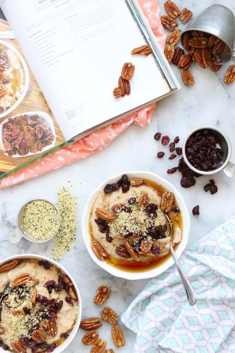 overhead shot of buckwheat porridge in white bowls on a marble surface with pecans, hempseed, cranberry garnishes scattered about