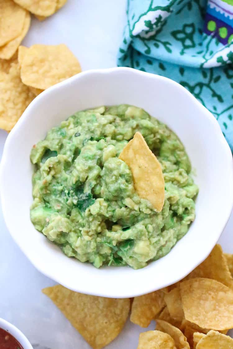 overhead closeup shot of guacamole in a white bowl with a tortilla chip sticking up and more chips nearby