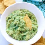 overhead shot of guacamole in a white bowl with corn chips surrounding and one stuck into the dip bowl