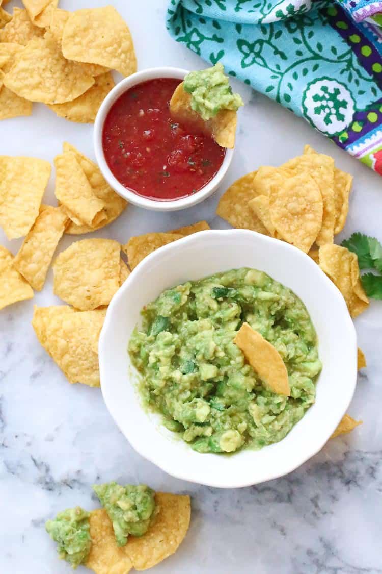 wide overhead shot of chips dipped into guacamole with salsa nearby, chips scattered around and colorful napkin in the corner