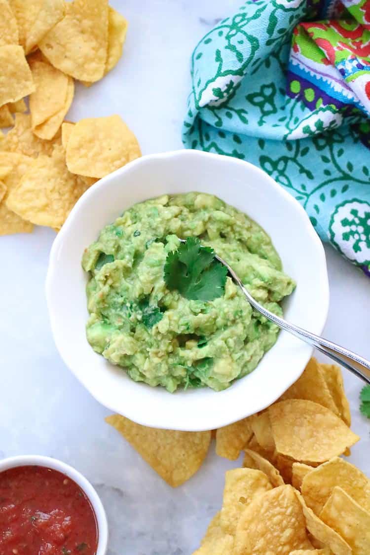 overhead shot of white bowl filled with guacamole with a garnish fo cilantro a ramekin of salsa nearby and tortilla chips laid out