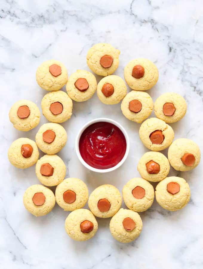 overhead shot of vegan corndogs arranged in a ring around ramekin full of ketchup on a marble surface