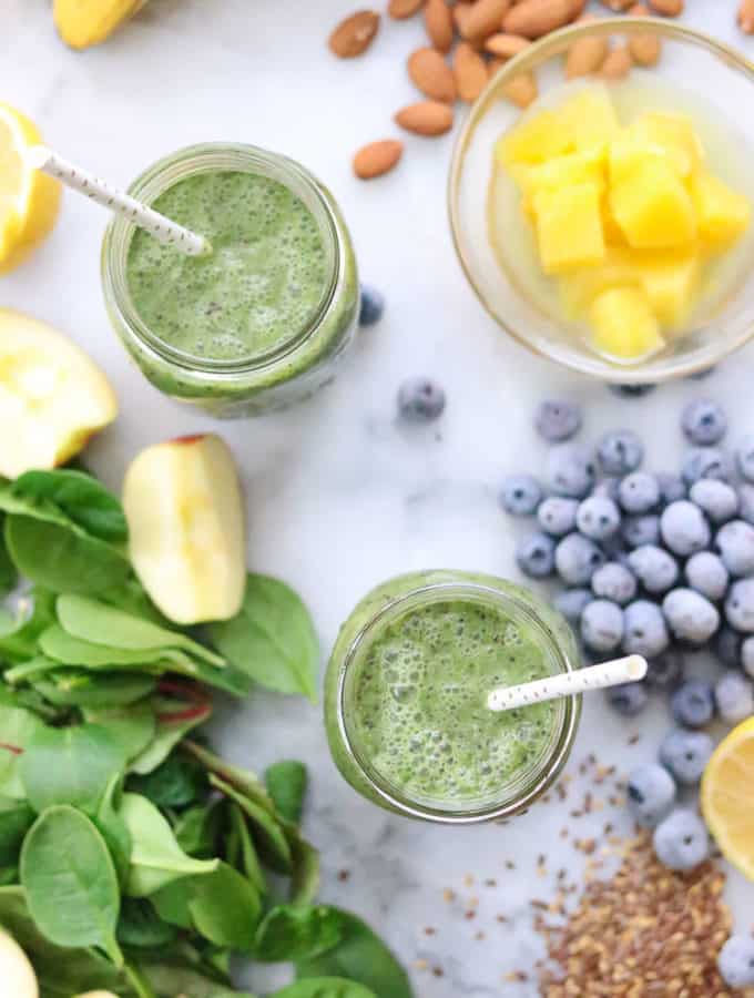 overhead shot of green smoothie in two mason jars with straws, surrounded by green smoothie ingredients
