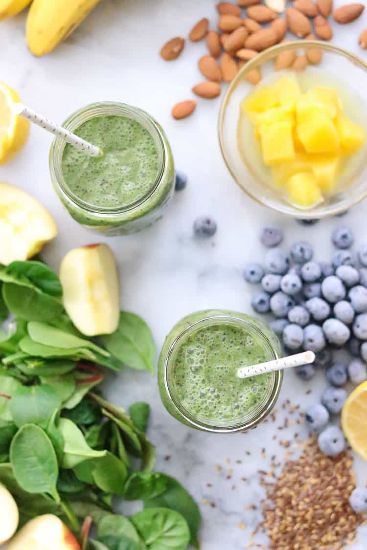 overhead shot of green smoothie in two mason jars with straws, surrounded by green smoothie ingredients