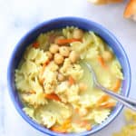 overhead shot of vegan chicken noodle soup in a blue bowl with bread and dried pasta nearby