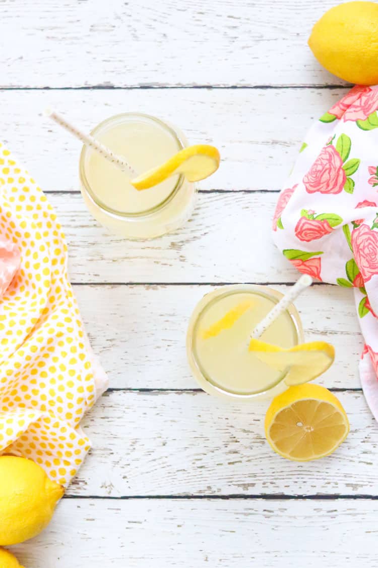overhead shot of two pint jars of lemon water on white wooden surface with two bright yellow and rose napkins and lemons around