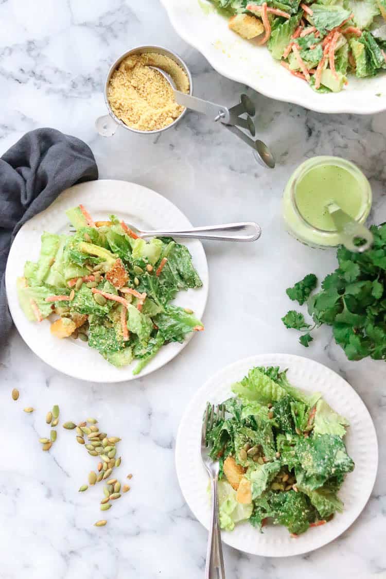 wide overhead shot of two plates of vegan caesar salad with jar of vegan caesar dressing beside and vegan parmesan in a cup nearby