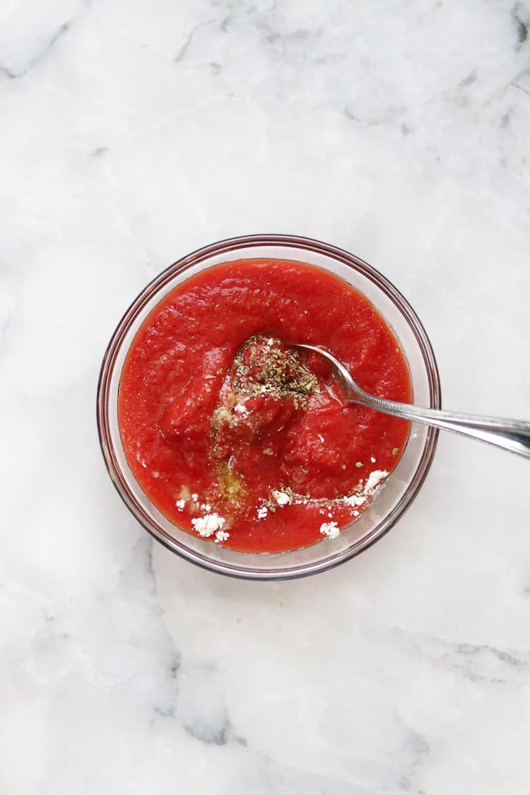 all ingredients for pizza sauce in a small clear bowl with spoon to mix