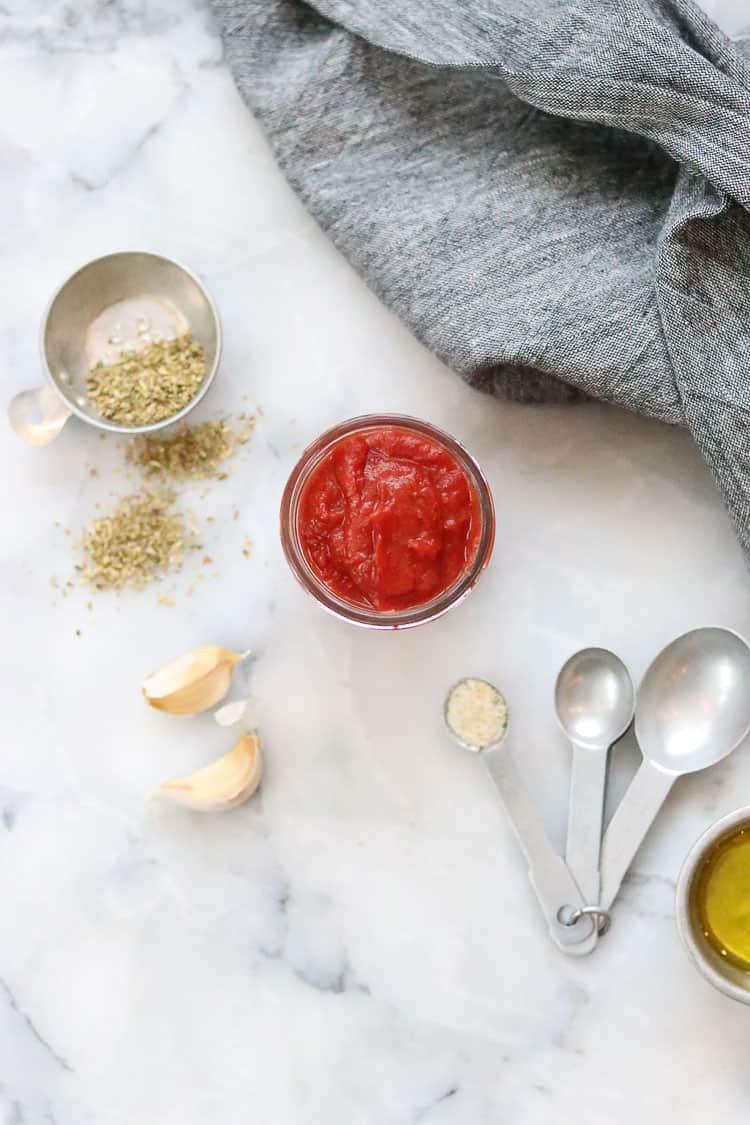 Overhead shot of pizza sauce in a small jar with measuring spoons and cups nearby filled with oregano, garlic salt and oil