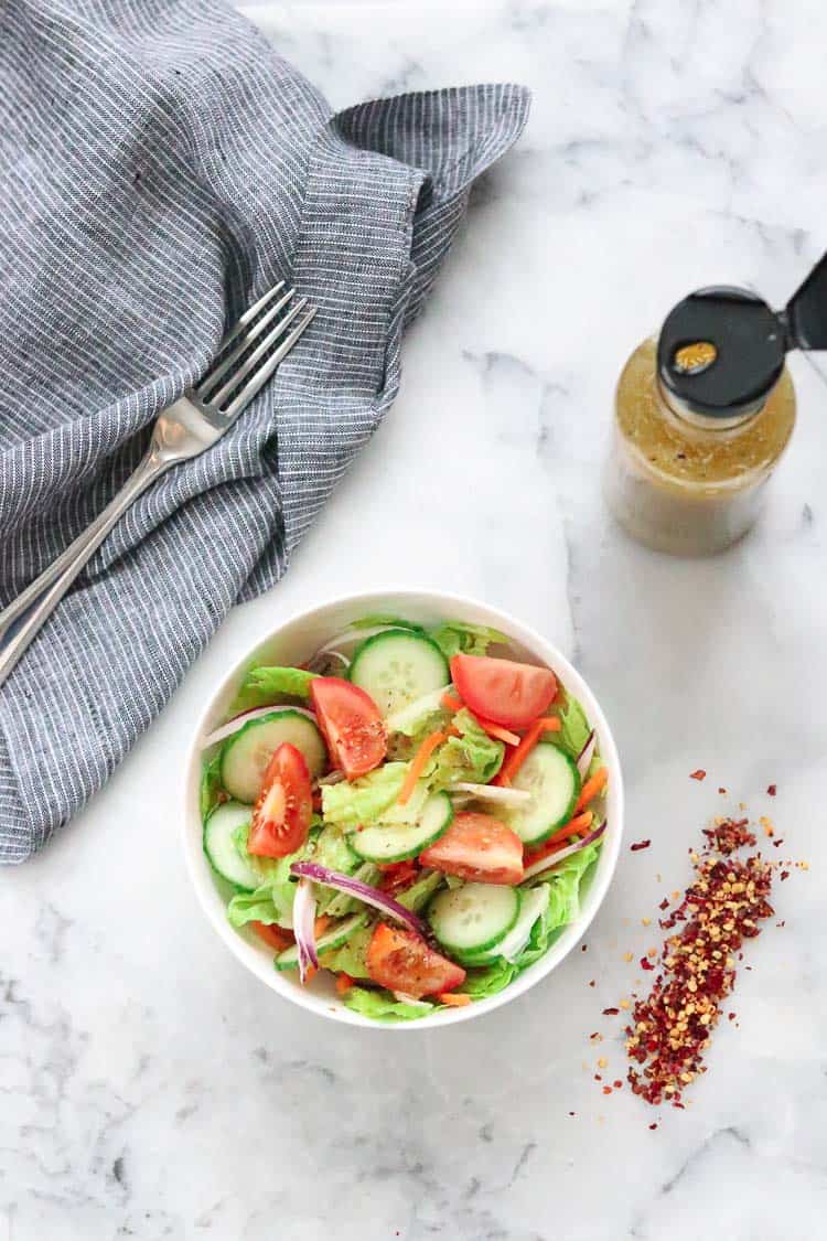 wide overhead shot of white bowl filled with salad with bottle of homemade Italian dressing in a bottle and red pepper scattered nearby