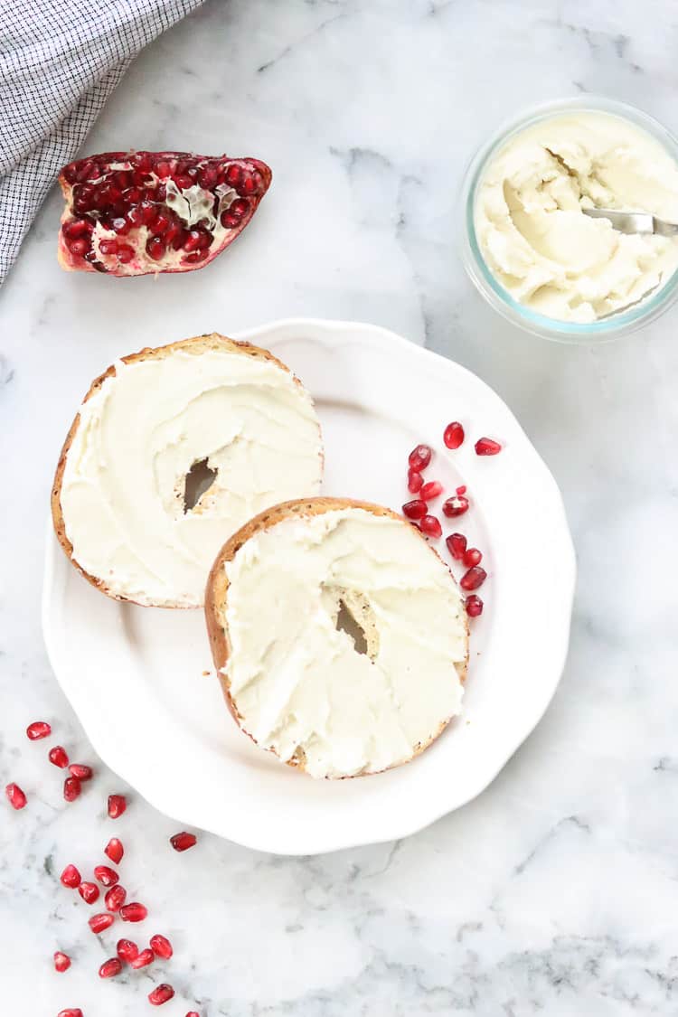 vegan cream cheese on bagel halves with pomegranates nearby 