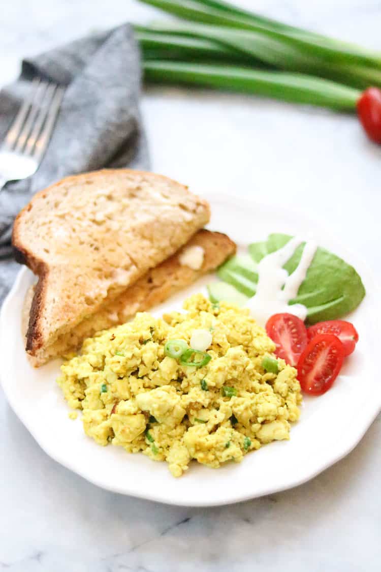 side overhead shot of tofu scramble on a white plate with sliced avocado and toast with napkin beside