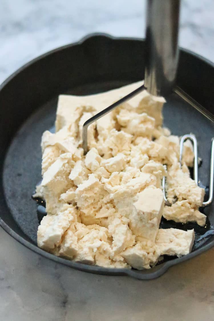 overhead shot of tofu being crumbled in black skillet with potato masher