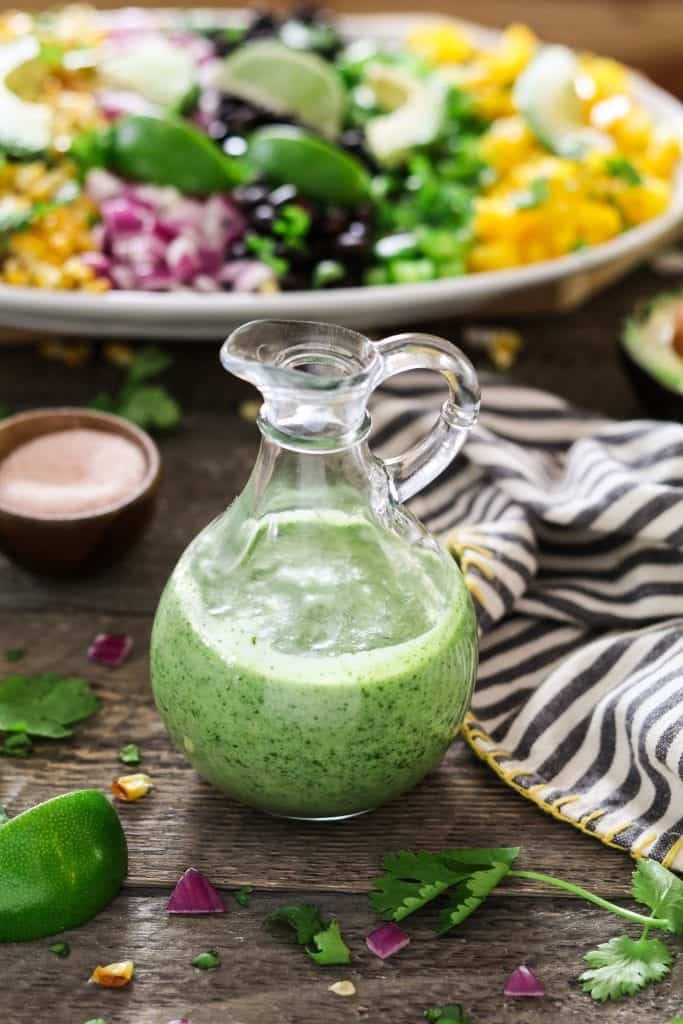 side overhead shot of green vegan dressing in a clear pitcher surrounded by red onion, lime, and cilantro pieces