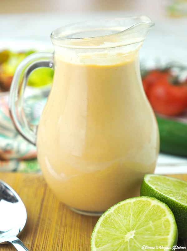 side shot of small pitcher of vegan dressing on wooden surface with limes in front