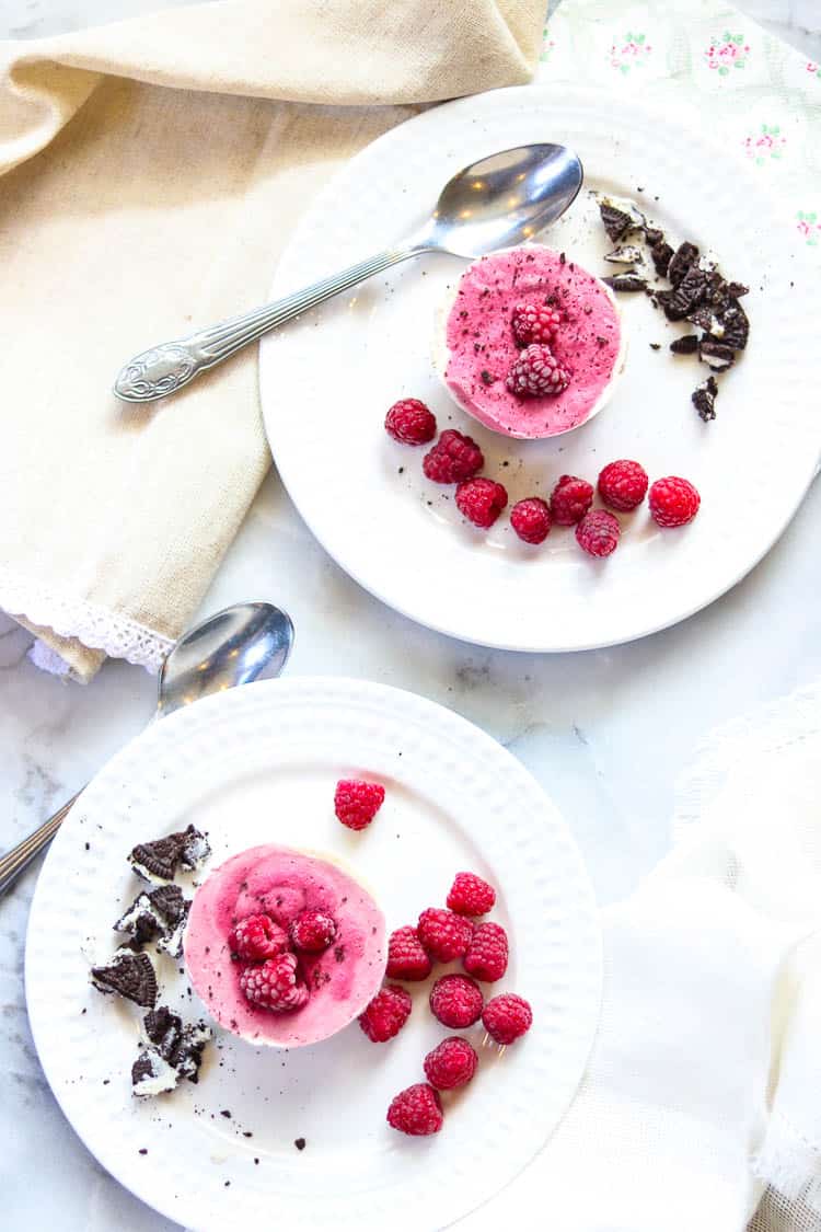 wide overhead shot of vegan ice cream cups being served on white plates with raspberries and crumbled chocolate cookies beside on white marble background
