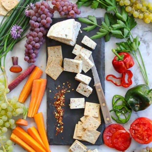 wide overhead shot of vegan cheese on a black slate board and surrounded by sliced veggies and fruit