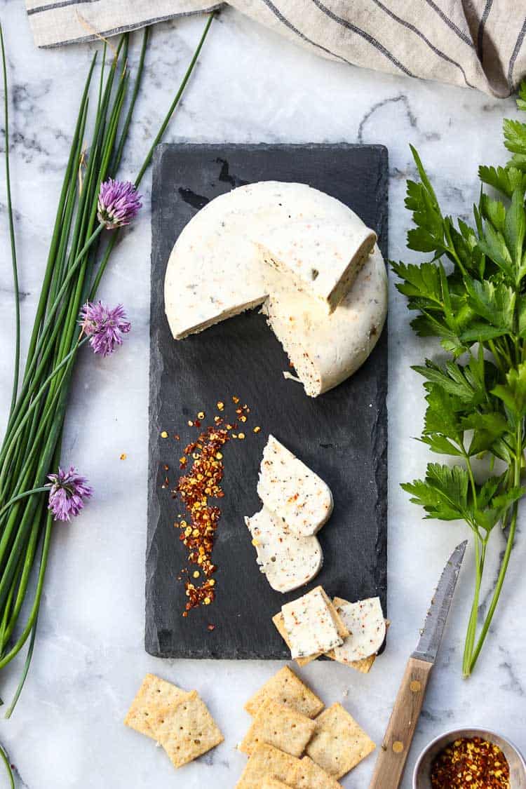 wide overhead shot of vegan herb cheese on a slate cheese board with crushed red pepper and fresh herbs nearby