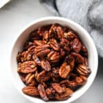 overhead shot of vegan candied pecans in a white bowl with a gray napkin and baking tray nearby