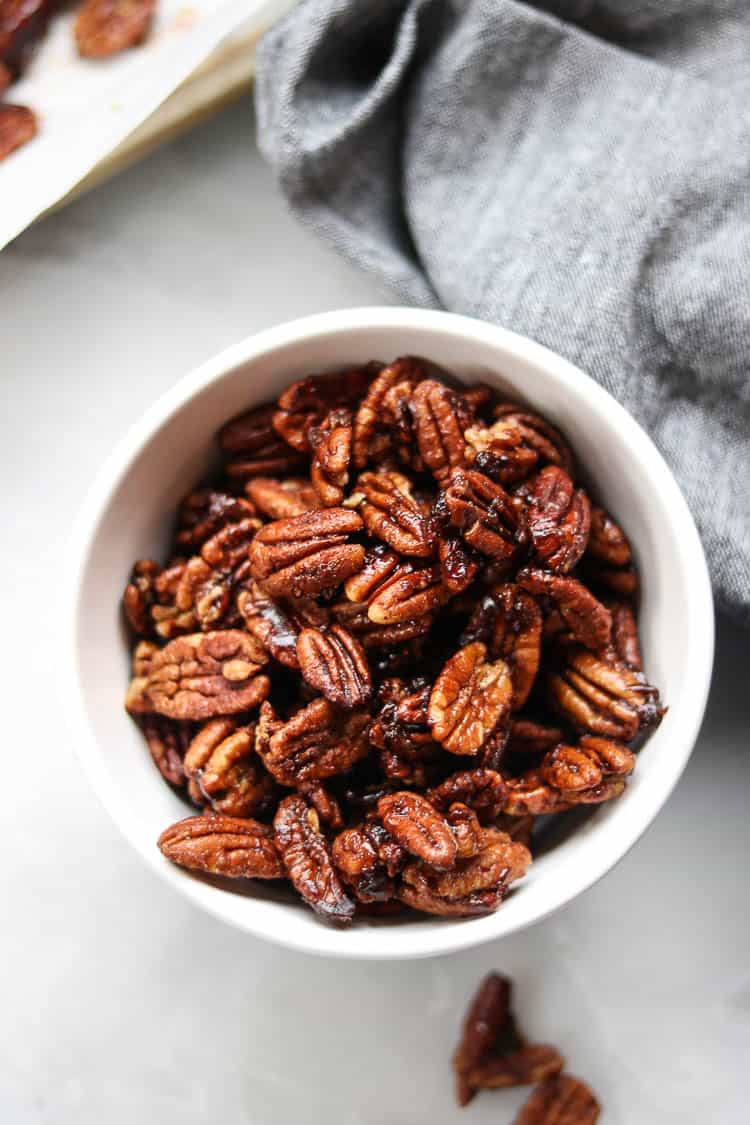 overhead shot of vegan candied pecans in a white bowl with a gray napkin and baking tray nearby