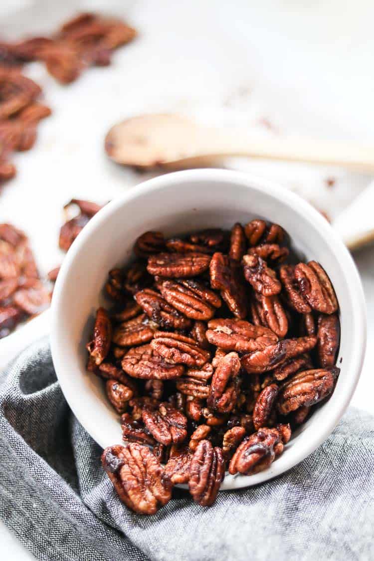 side view of white bowl of candied pecans pouring out on a gray napkin